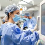 Diverse Team of Professional Surgeons Performing Invasive Surgery on a Patient in the Hospital Operating Room. Nurse Hands Out Instruments to surgeon, Anesthesiologist Monitors Vitals.